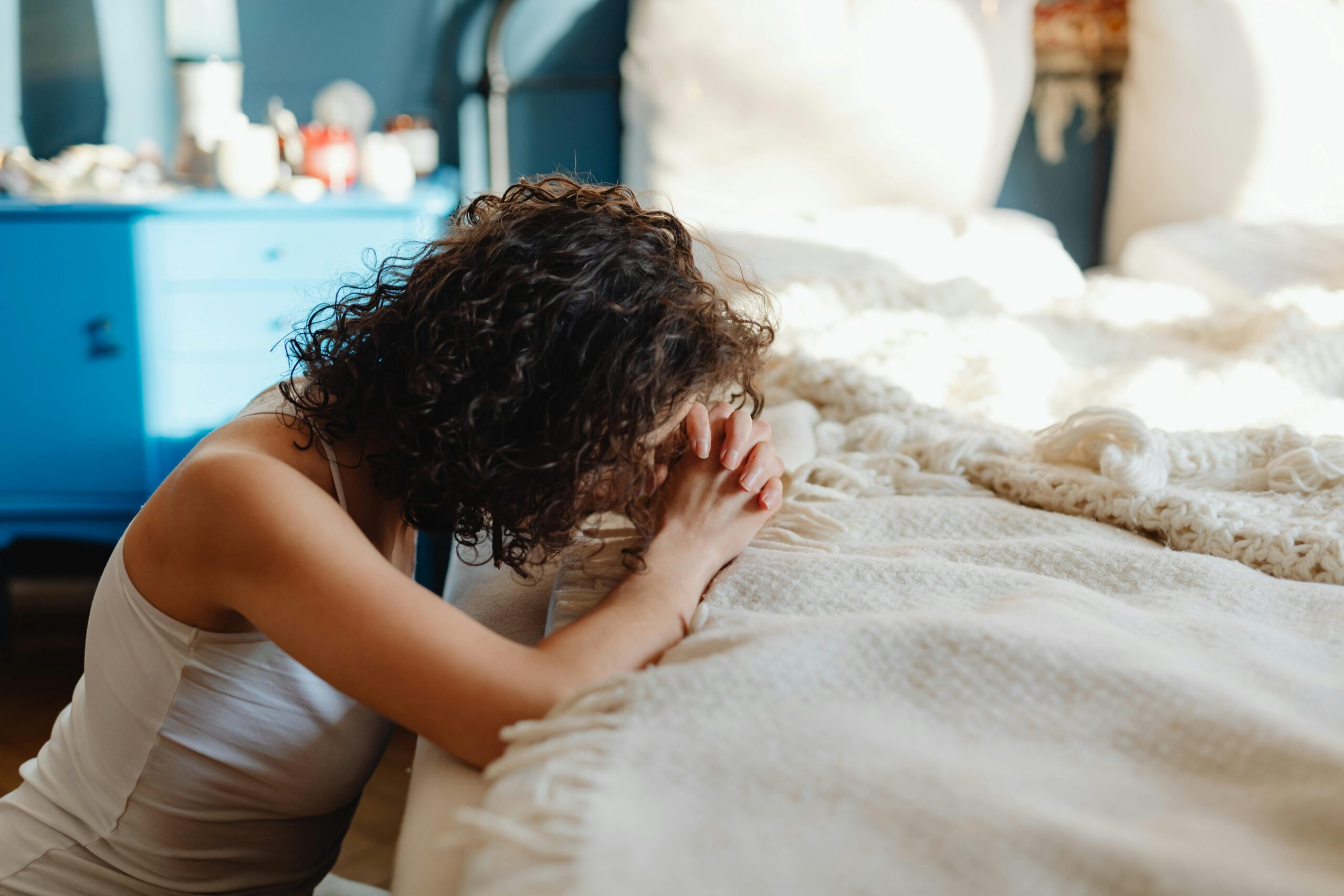 Woman sitting on the floor beside her bed, hands clasped in a moment of emotional vulnerability.