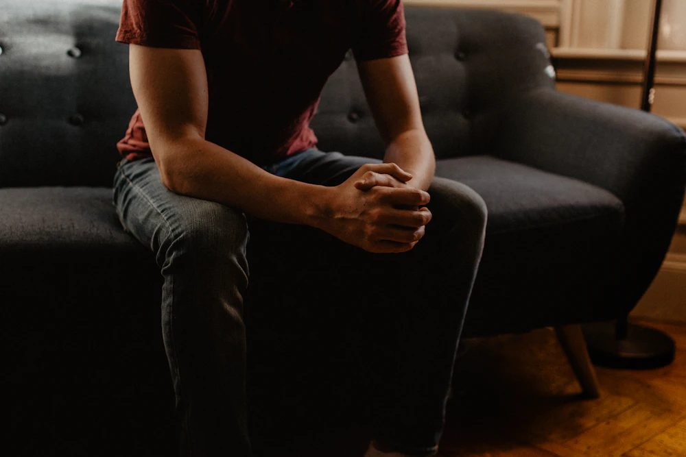 An adult seated on a sofa with hands clasped and face outside the frame
